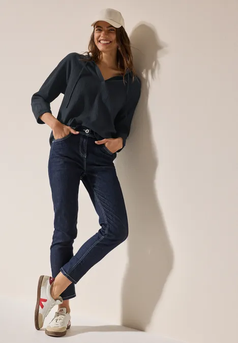 Frau in dunkler Jeans, Pullover und Cap lehnt lächelnd an einer Wand.